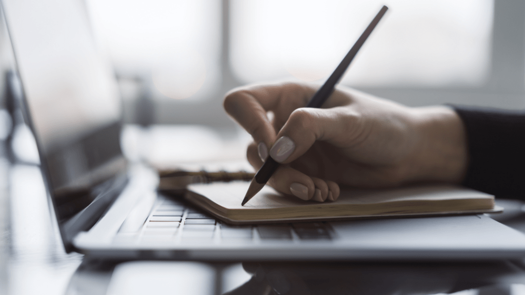 A woman writing on a notepad placed on top of a laptop