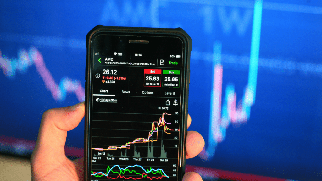 a person holding a smart phone in front of a stock market graph